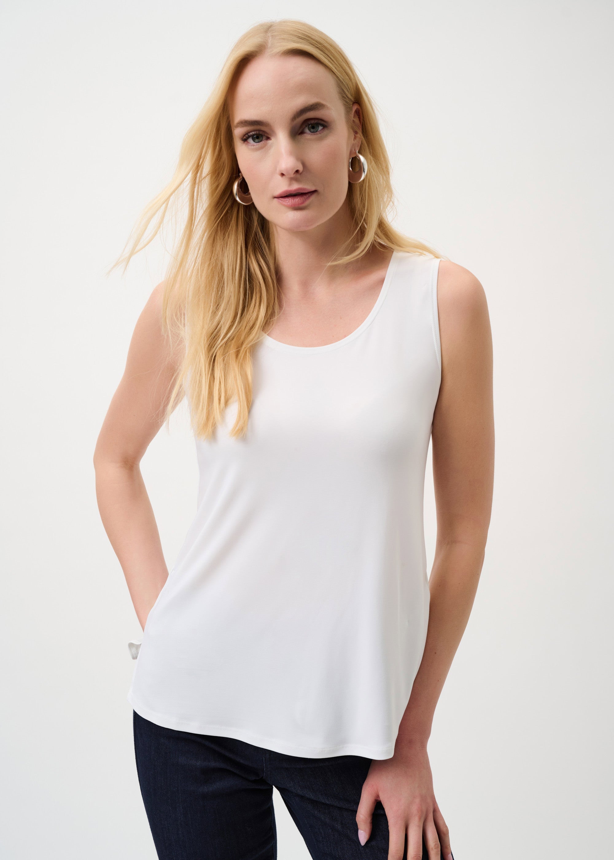 Woman wearing a white sleeveless top and dark pants on a plain background