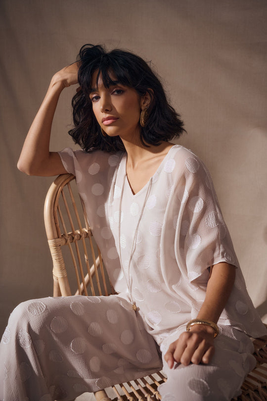 Light colored chiffon poncho top with polka dot pattern worn by model seated on wicker chair