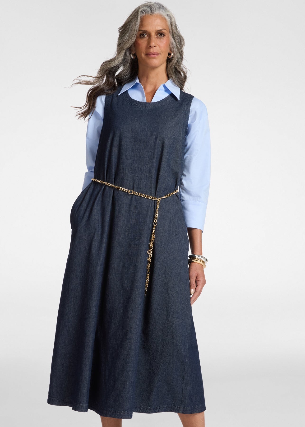 Woman wearing a navy pinafore dress over a light blue shirt on a white background