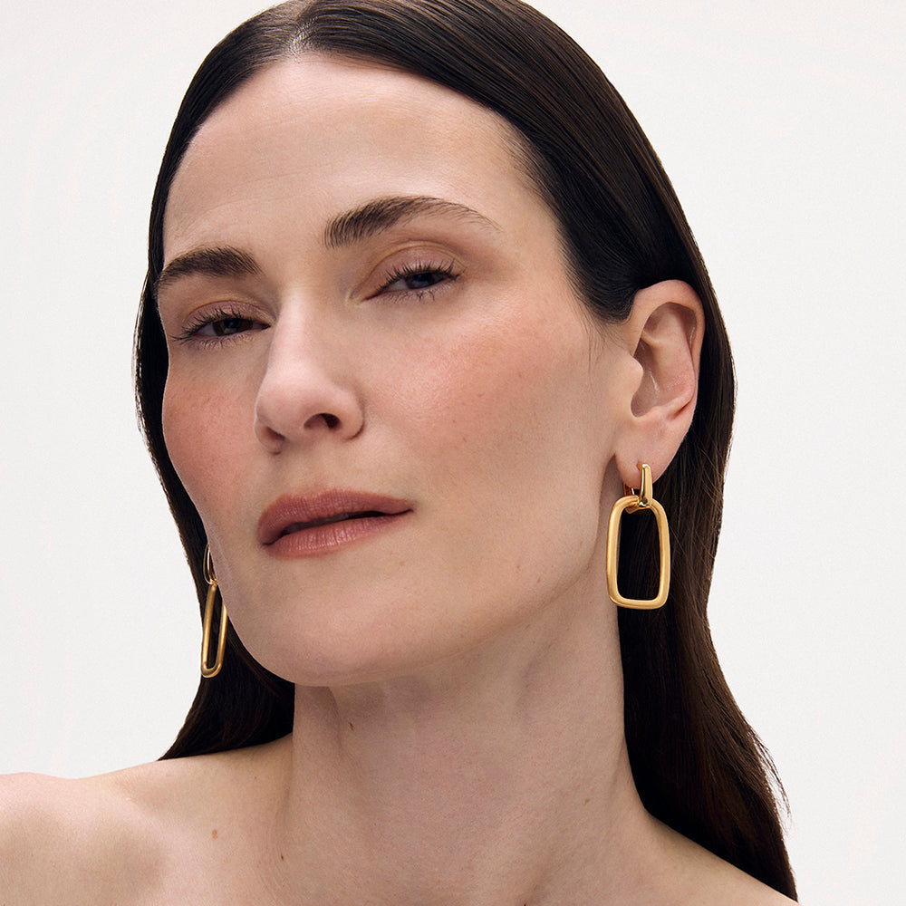 Model wearing gold geometric drop hoop earrings with rectangular open-frame pendant