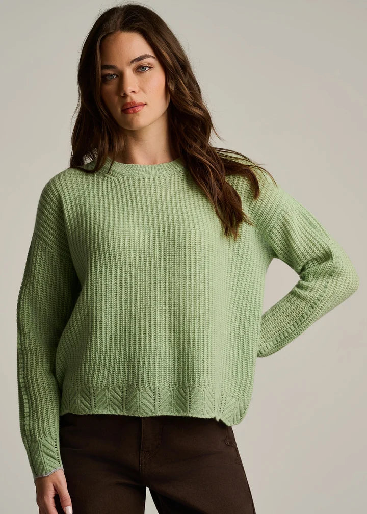 A woman wearing a light sage green ribbed knit sweater and dark brown pants, posing against a neutral light gray studio background.