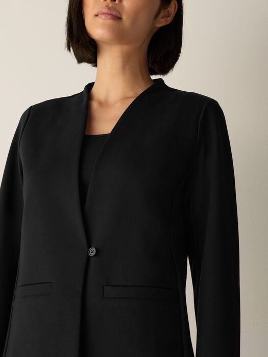 Collarless black blazer with single button closure and welt pockets, front detail view on model