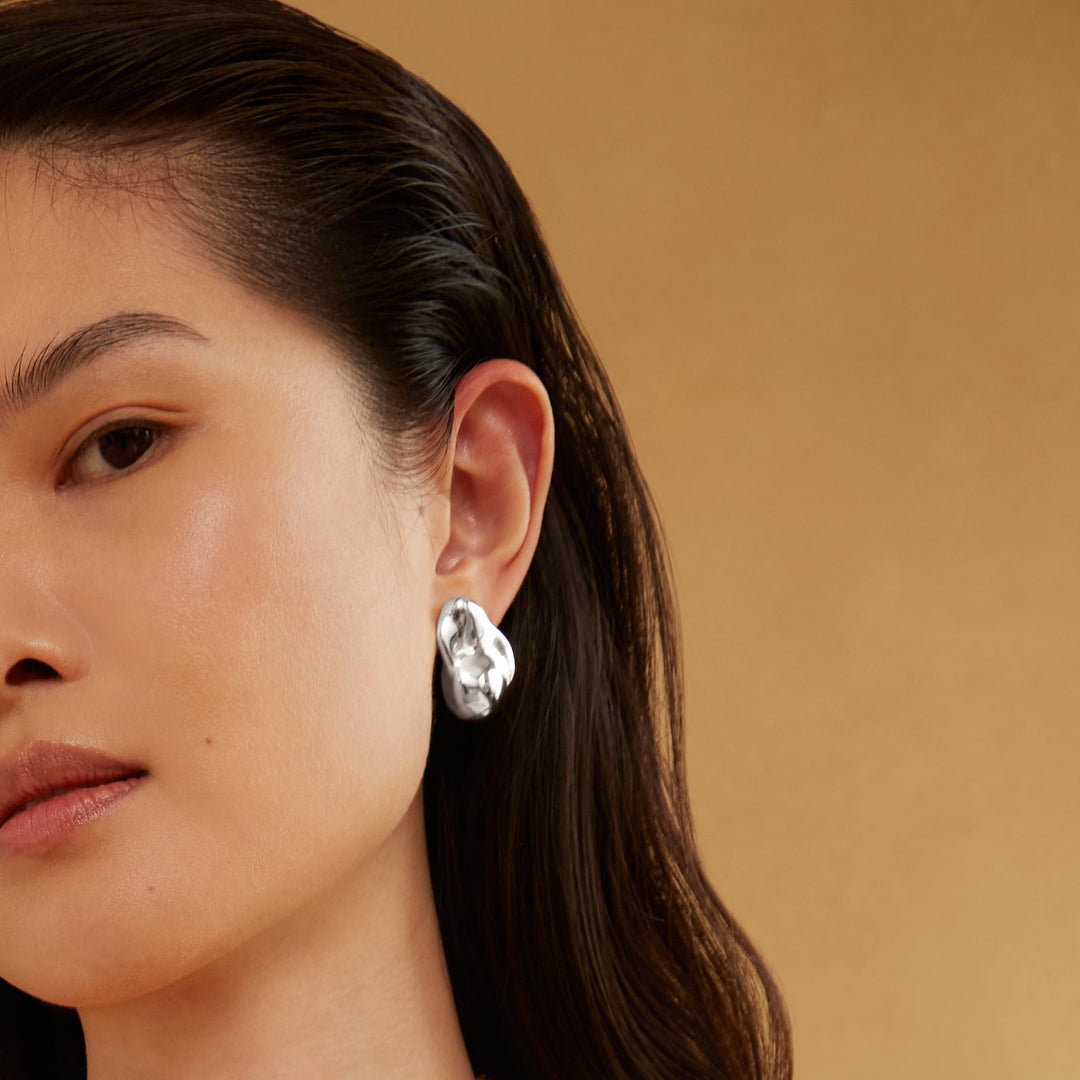 Silver molten-style stud earring worn on model's ear, irregular organic shape