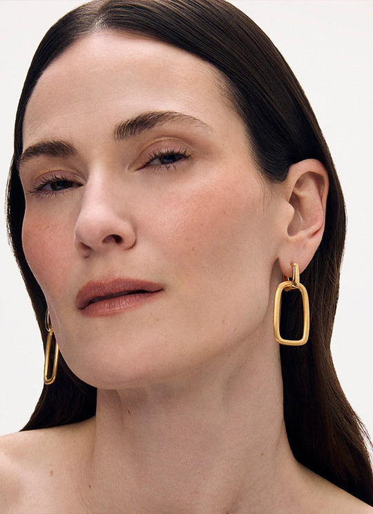 Model wearing gold drop hoop earrings with open rectangular pendant, close-up view